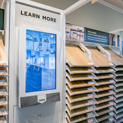 A sample display stand with a touchscreen showing a kitchen interior, next to a rack of wood-look flooring samples.